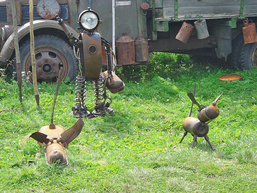 Photographie d'un jardin avec des objets en métal recyclé représentant une personne et deux chiens en train de jouer. La personne pourrait être un vétérinaire.